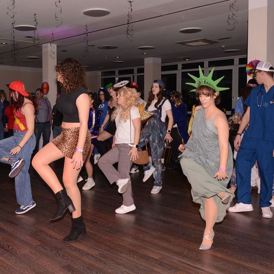 Eine Gruppe von Menschen in Kostümen tanzt auf einem Holzboden bei einer Veranstaltung. Einige tragen Hüte und Stiefel, und eine trägt ein Statue of Liberty-Kostüm.