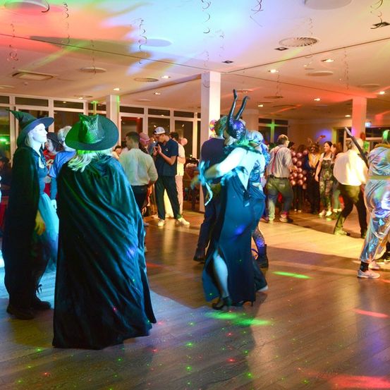 Eine Tanzfläche mit Menschen in Kostümen. Einige tanzen, andere stehen und schauen zu, während bunte Lichter den Raum erhellen.