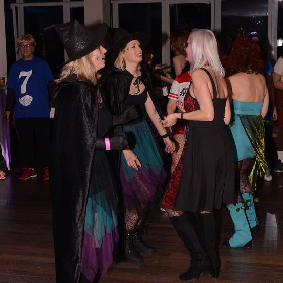 Mehrere Erwachsene in Halloween-Kostümen tanzen auf einem Holzboden. Eine Frau trägt einen Hexenhut und einen schwarzen Umhang. Eine andere Frau trägt ein schwarzes Kleid und Stiefel.