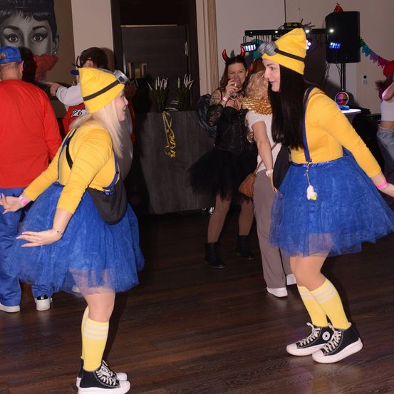 Zwei Frauen in Minions-Kostümen tanzen auf einem Holzboden bei einer Party. Eine Frau in einem schwarzen Kleid und andere sind im Hintergrund.