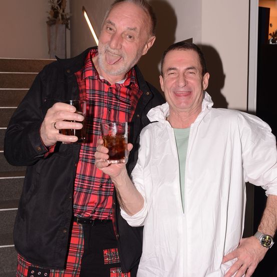 Zwei Männer stehen zusammen, einer trägt ein kariertes Hemd und hält ein Getränk, der andere ein weißes Laborkittel mit einem Getränk in den Händen, beide lächeln.