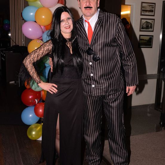 Eine Frau und ein Mann in Kostümen stehen nebeneinander und lächeln vor einem Hintergrund bunter Luftballons.