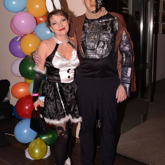 Ein Mann und eine Frau in Kostümen stehen vor einem bunten Luftballon-Hintergrund. Die Frau trägt ein schwarzes und weißes Kleid mit einem weißen Band, während der Mann als Roboter gekleidet ist.