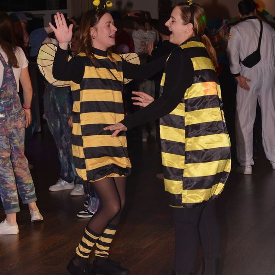 Zwei Frauen in Bienenkostümen tanzen in einem Raum mit anderen Leuten. Eine Frau hat ihre Hände erhoben, während die andere lächelt.