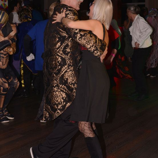 Ein Paar tanzt eng auf einem Holzboden bei einer Kostümparty. Die Frau trägt ein schwarzes Kleid und der Mann einen goldgemusterten Anzug. Andere Partygäste in Kostümen schauen zu.