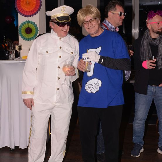 Zwei Leute in Kostümen stehen bei einer Party. Einer hält ein Getränk und trägt eine Matrosenuniform, der andere hält einen Becher und trägt ein blaues T-Shirt. Dahinter stehen zwei weitere Leute, einer in einem Anzug und der andere in Freizeitkleidung.