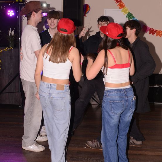 Eine Gruppe junger Leute in Freizeitkleidung tanzt auf einem Holzboden. Zwei Frauen tragen rote Mützen, während andere um sie herum stehen. Ein Lautsprecher und bunte Dekorationen sind im Hintergrund zu sehen.