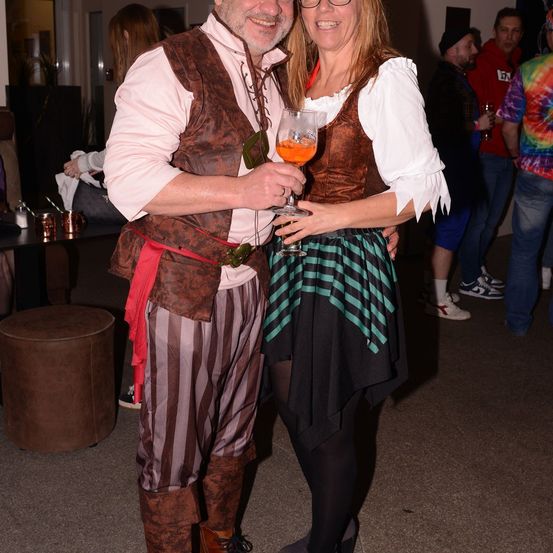 Ein Paar, als Piraten gekleidet, steht lächelnd zusammen mit Getränken in der Hand. Sie sind von anderen auf einer Party umgeben, mit Tischen und Stühlen im Hintergrund.