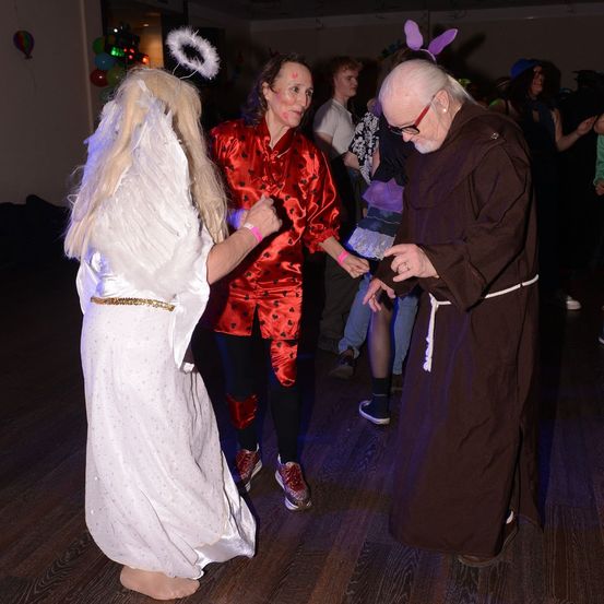 Zwei Menschen in Kostümen tanzen auf einem Holzboden, ein dritter in einem Mönchsgewand steht in der Nähe. Hinter ihnen tanzen weitere Menschen. An der Wand hängen Luftballons und eine Heißluftballon-Dekoration.