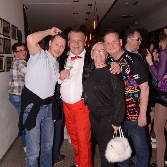 Eine Gruppe von Menschen steht in einem Saal zusammen, ein Mann hält ein Weinglas und eine Frau eine weiße Handtasche. Hinter ihnen stehen andere, möglicherweise bei einer Veranstaltung.