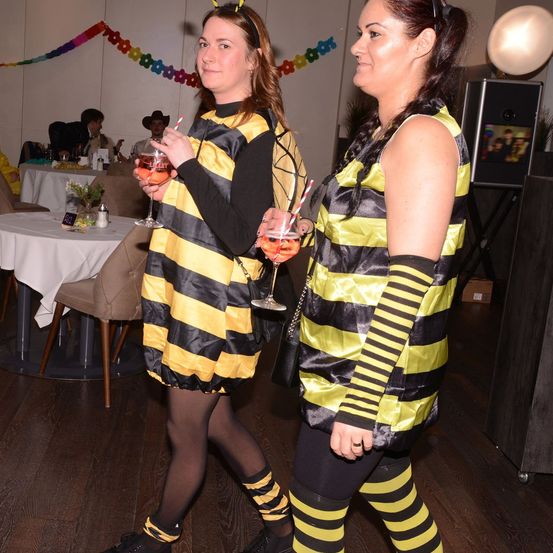 Zwei Frauen in Bienenkostümen gehen mit Getränken in einem dekorierten Raum. Eine hält ein Weinglas und die andere einen Cocktail. Ein Tisch und Stühle sind hinter ihnen, und ein Lautsprecher befindet sich rechts.