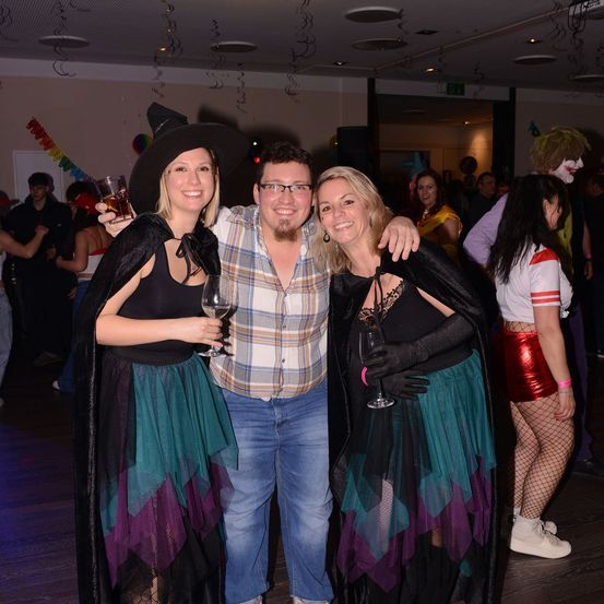 Drei Personen posieren für ein Foto auf einer Party. Zwei Frauen in Hexenkostümen und ein Mann in Freizeitkleidung. Alle halten Getränke. Im Hintergrund sind weitere Menschen in verschiedenen Kostümen zu sehen, mit Dekorationen, die von der Decke hängen.