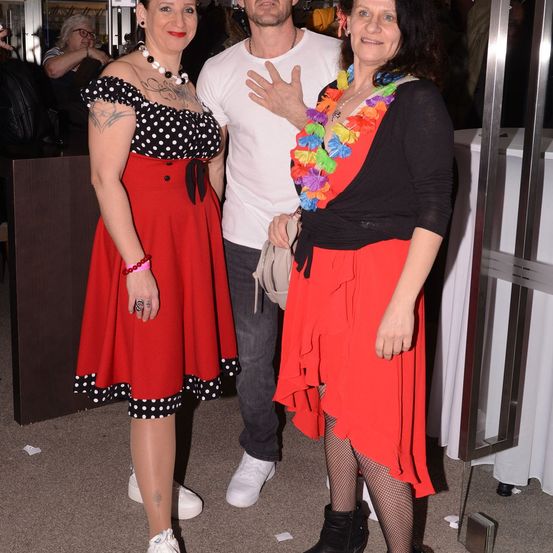 Drei Personen bei einer Veranstaltung. Zwei Frauen in roten Kleidern und ein Mann in einem weißen T-Shirt. Sie lächeln und posieren für ein Foto.