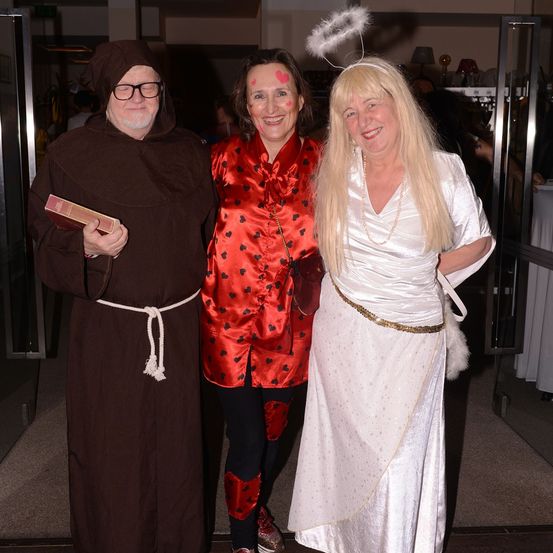 Drei Erwachsene in Kostümen posieren für ein Foto in einem Raum. Ein Mann hält ein Buch, eine Frau trägt ein rotes Herzmuster-Outfit und eine andere Frau trägt ein weißes Engelkostüm.