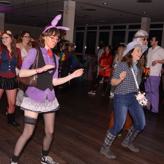 Eine Gruppe von Menschen in Kostümen tanzt in einem Raum. Eine Frau mit Hasenohren führt die Gruppe an. Eine andere Frau hält ein Bier und eine Kuscheltier. Der Raum hat Holzböden und Glasfenster.