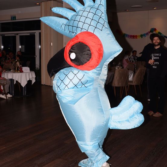 Eine Person in einem blauen Papageienkostüm steht in einem Raum mit Holzböden. Hinter ihnen sitzen Leute an Tischen. Ein Mann hält ein Glas Wein.