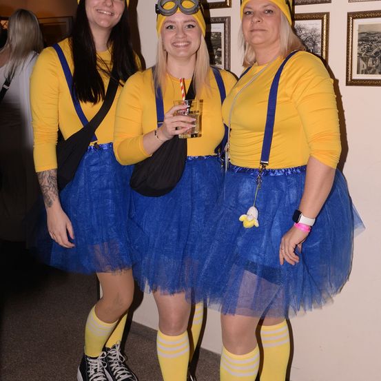 Drei Frauen in gelben Minion-Kostümen mit blauen Tüllröcken und Schutzbrillen posieren für ein Foto, eine hält ein Getränk in der Hand.