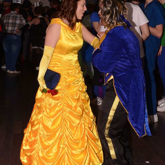 Eine Frau in einem gelben Belle-Kostüm tanzt mit einem Mann in einem blauen Biest-Kostüm auf einer Kostümparty. Sie sind von anderen Menschen umgeben.