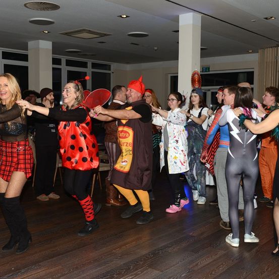 Eine Gruppe von Menschen in Kostümen tanzt in einem Raum. Eine Person trägt einen roten Hut. Der Raum hat einen Holzboden und Glasfenster.