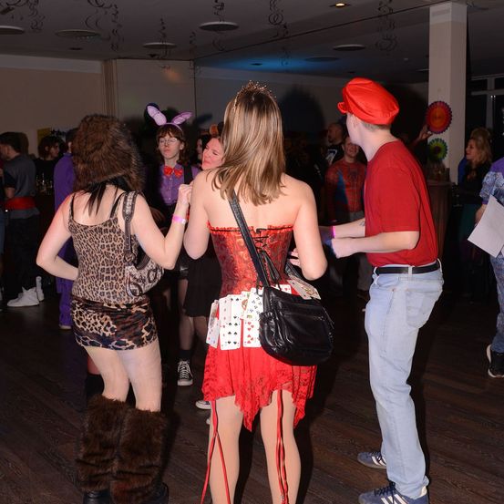 Eine Gruppe von Menschen in einem Raum, eine Frau trägt eine Krone, eine andere ein Leopardenkleid, und ein Mann in einem roten Hut. Der Raum hat Holzböden und eine festliche Atmosphäre.