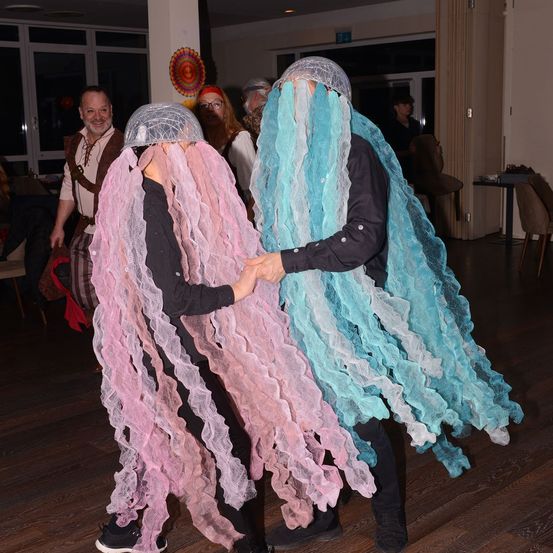 Zwei Menschen in rosa und blauen Quallenkostümen tanzen in einem Saal. Andere Menschen in verschiedenen Kostümen sind im Hintergrund.