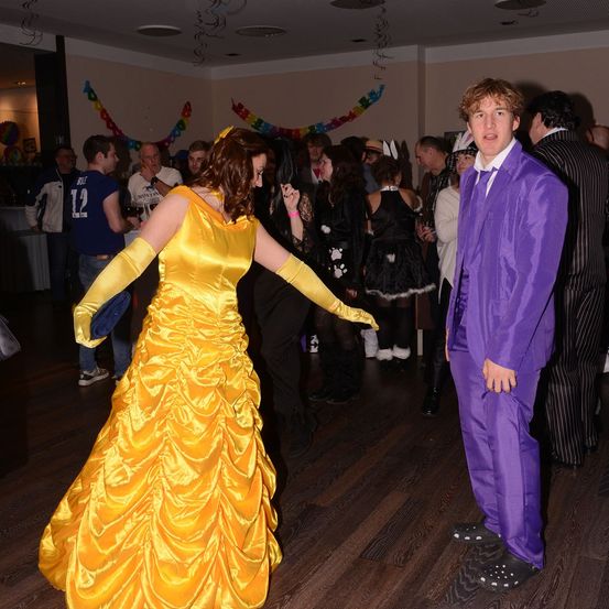 Eine Frau in einem gelben Belle-Kostüm aus Die Schöne und das Biest tanzt in einem überfüllten Raum mit anderen in Kostümen. Ein Mann in einem lila Anzug steht in der Nähe.