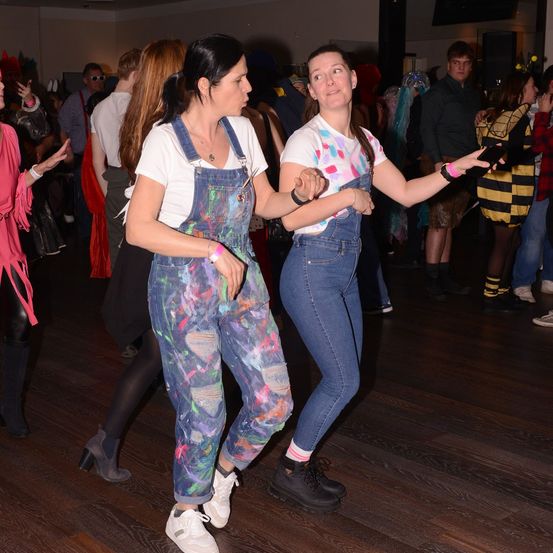 Zwei Frauen tanzen zusammen auf einem Holzboden, in Overalls gekleidet. Eine Menschenmenge in Kostümen umgibt sie.
