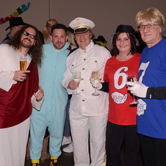 Eine Gruppe von Menschen in verschiedenen Kostümen posiert für ein Foto bei einer Veranstaltung und hält Weingläser in den Händen. Einer trägt eine Kapitänsmütze, ein anderer ein blaues Sweatshirt und ein dritter ein rotes T-Shirt mit der Nummer 6.