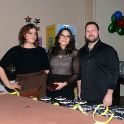 Three people stand behind a counter with glasses, smiling for a photo. The woman in the middle wears a blue headband, the woman on the left has a star headband. The man wears a black shirt. Behind them is a yellow banner with green balloons.