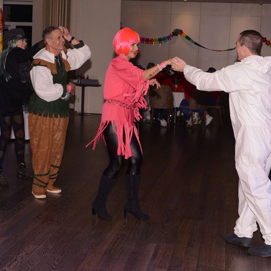 Eine Gruppe von Menschen in Kostümen tanzt auf einem Holzboden. Eine Frau in Pink winkt, während zwei Männer ihre Hände halten.