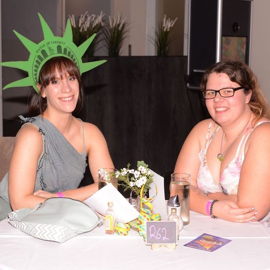 Zwei Frauen sitzen an einem Tisch, eine trägt eine Statue of Liberty-Kopfbedeckung und lächelt. Auf dem Tisch steht eine Blumenarrangement, eine Handtasche und ein Schild mit der Aufschrift RG2.
