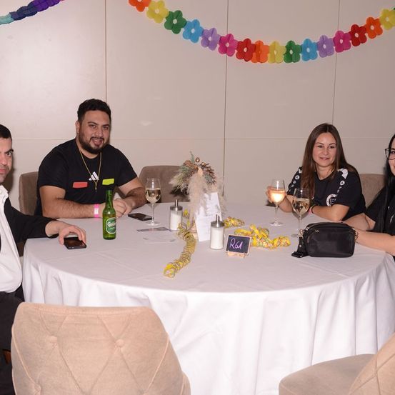 Fünf Personen sitzen bei einer Party um einen Tisch. Vier von ihnen halten Weingläser in den Händen, während die fünfte ein Handy hält. Eine grüne Flasche steht auf dem Tisch. Eine bunte Girlande hängt darüber.