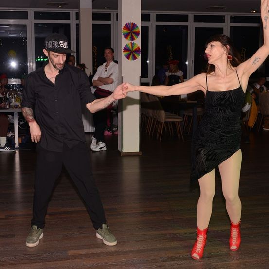 Ein Mann und eine Frau tanzen bei einer lebhaften Veranstaltung, mit Dekorationen im Hintergrund und Menschen, die an Tischen sitzen.