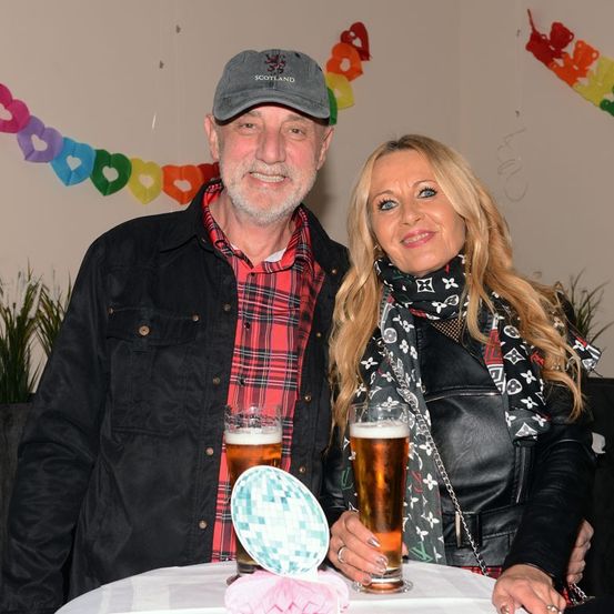 Ein lächelndes Paar posiert für ein Foto mit Biergläsern. Sie stehen bei einem Tisch mit dekorativen Papierbällen. Dahinter eine Wand mit bunten Papierherzen und einer Topfpflanze.