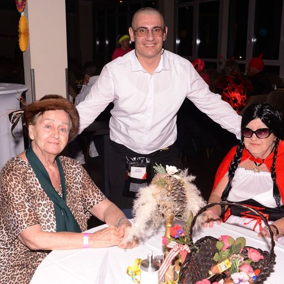 Drei Menschen sitzen an einem Tisch, ein Mann steht hinter ihnen. Eine Frau in Leopardenmuster und eine Frau in Rot mit einem Korb mit Blumen.