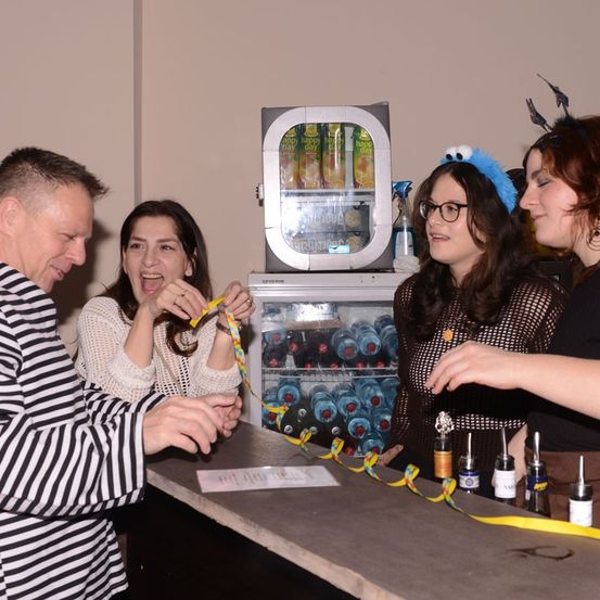 Eine Gruppe von Menschen steht um einen Tresen und lacht. Eine Frau in einer weißen Jacke hält eine gelbe Schleife. Dahinter steht ein Kühlschrank mit Getränken, und eine Frau mit einer blauen Haube lächelt.