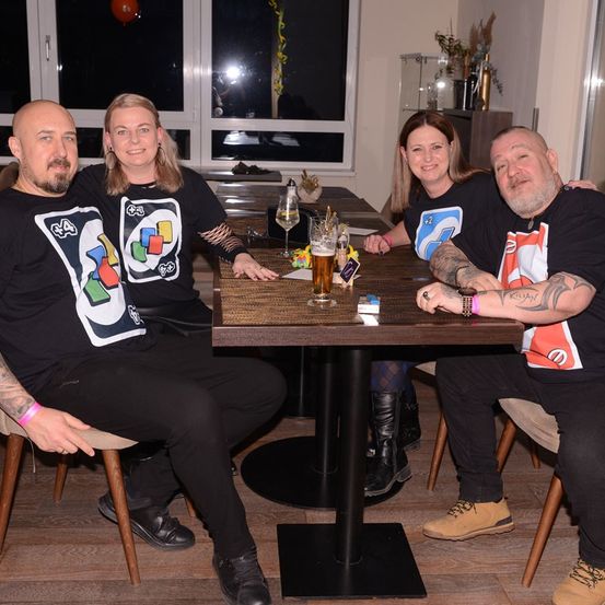 Vier Personen in passenden T-Shirts sitzen um einen Tisch mit Getränken und lächeln. Einer hat eine Tätowierung am Arm. Im Hintergrund ist ein Fenster mit einer Reflexion zu sehen.