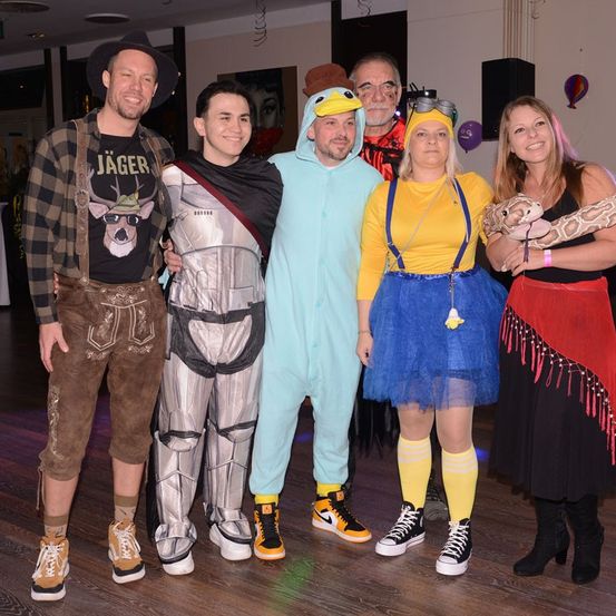 Sechs Menschen in Kostümen stehen zusammen auf einem Holzboden. Einer hält eine Schlange. Einer trägt ein Roboterkostüm. Einer trägt einen grünen Hut und eine Entenmaske.