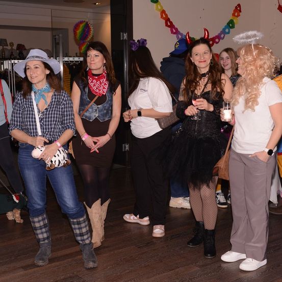 Frauen in verschiedenen Kostümen stehen in einem Raum, möglicherweise auf einer Party. Die Frau auf der linken Seite hält ein Plüschtier. Eine Frau auf der rechten Seite trägt ein weißes Shirt und eine blonde Perücke.