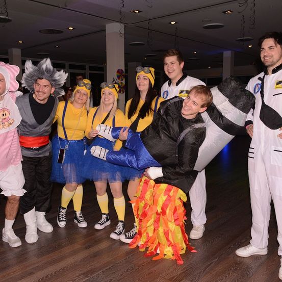 Eine Gruppe von Menschen in verschiedenen Kostümen posiert für ein Foto. Ein Mann in einem schwarzen und weißen Kostüm liegt mit erhobenen Beinen auf dem Boden.