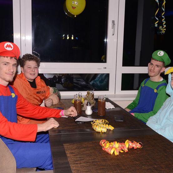 Vier Personen in Mario-Kostümen sitzen um einen Tisch, mit Getränken und Dekorationen. Ein großes Fenster dahinter spiegelt den Nachthimmel wider.