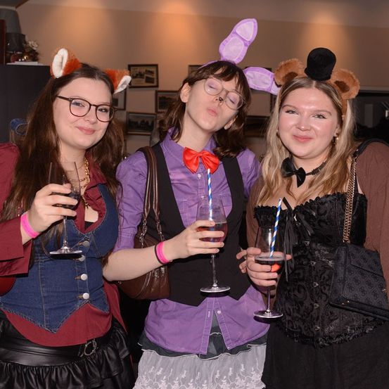 Drei Frauen in Kostümen posieren für ein Foto. Eine trägt ein Katzenkostüm, eine andere trägt eine Brille und ein lila Hemd, und die dritte hat eine Fliege und einen pelzigen Hut. Sie halten Gläser mit Wein.