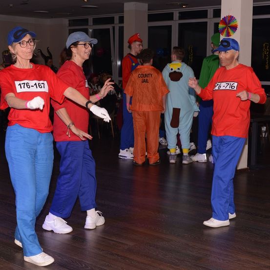Eine Gruppe von Erwachsenen in roten T-Shirts mit Nummern und blauen Hosen tanzt. Einige Personen tragen Kostüme.