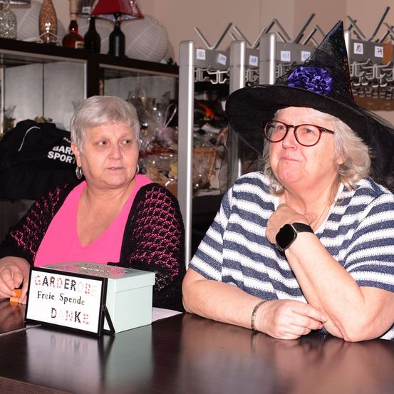 Zwei Frauen sitzen an einem Tresen, eine mit einem Hexenhut, beide lächeln. Eine Frau hält eine Box mit 'GARDERE FREIE SPENDE' darauf. Dahinter ein Regal mit Flaschen und einem schwarzen Kleidungsstück.