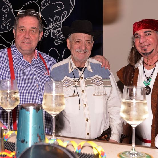 Drei Männer stehen an einer Bar mit Weingläsern. Der Mann links trägt Hosenträger, der Mann in der Mitte einen Cowboyhut und der Mann rechts eine rote Bandana.