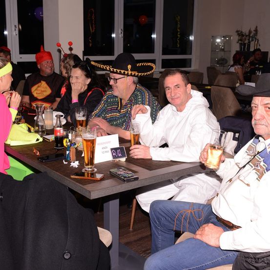 Eine Gruppe von Menschen sitzt um einen Tisch herum und trägt verschiedene Kostüme. Sie halten Getränke in den Händen. Einige tragen Hüte. Ein Mann auf der linken Seite trägt eine pinke Stirnband.