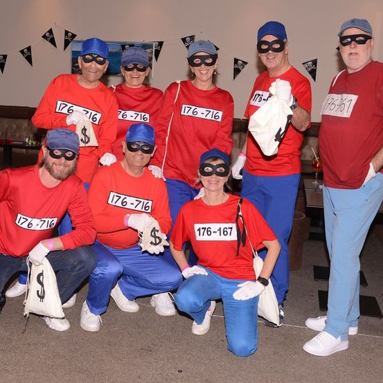 Eine Gruppe von Menschen, die rote Lätzchen, blaue Hosen und Masken tragen, posiert für ein Foto. Sie stehen und knien auf einem Teppichboden. Sie haben Nummern auf ihren Lätzchen, und einige von ihnen halten weiße Tüten.