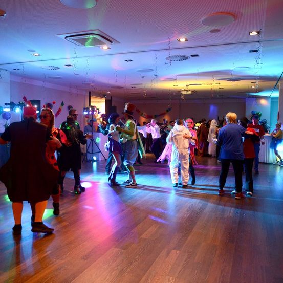 Ein Saal voller Menschen in Kostümen, die tanzen und von mehrfarbigen Lichtern beleuchtet werden.