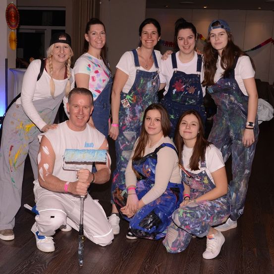 Eine Gruppe von Menschen, darunter Frauen und ein Mann, sind in bemalte Latzhosen gekleidet. Sie posieren für ein Foto mit einem lächelnden Mann, der einen Farbroller hält. Der Raum ist schwach beleuchtet, und es gibt Dekorationen im Hintergrund.
