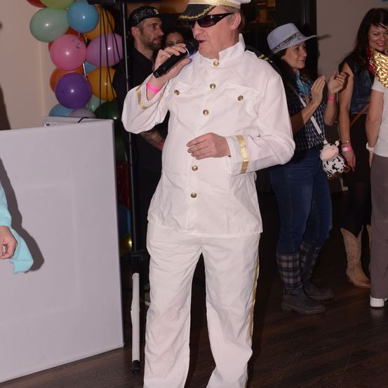Ein Mann in einem weißen Uniform mit goldenen Akzenten und einem Hut singt in ein Mikrofon auf einer Tanzfläche. Hinter ihm jubeln Menschen und halten Luftballons.